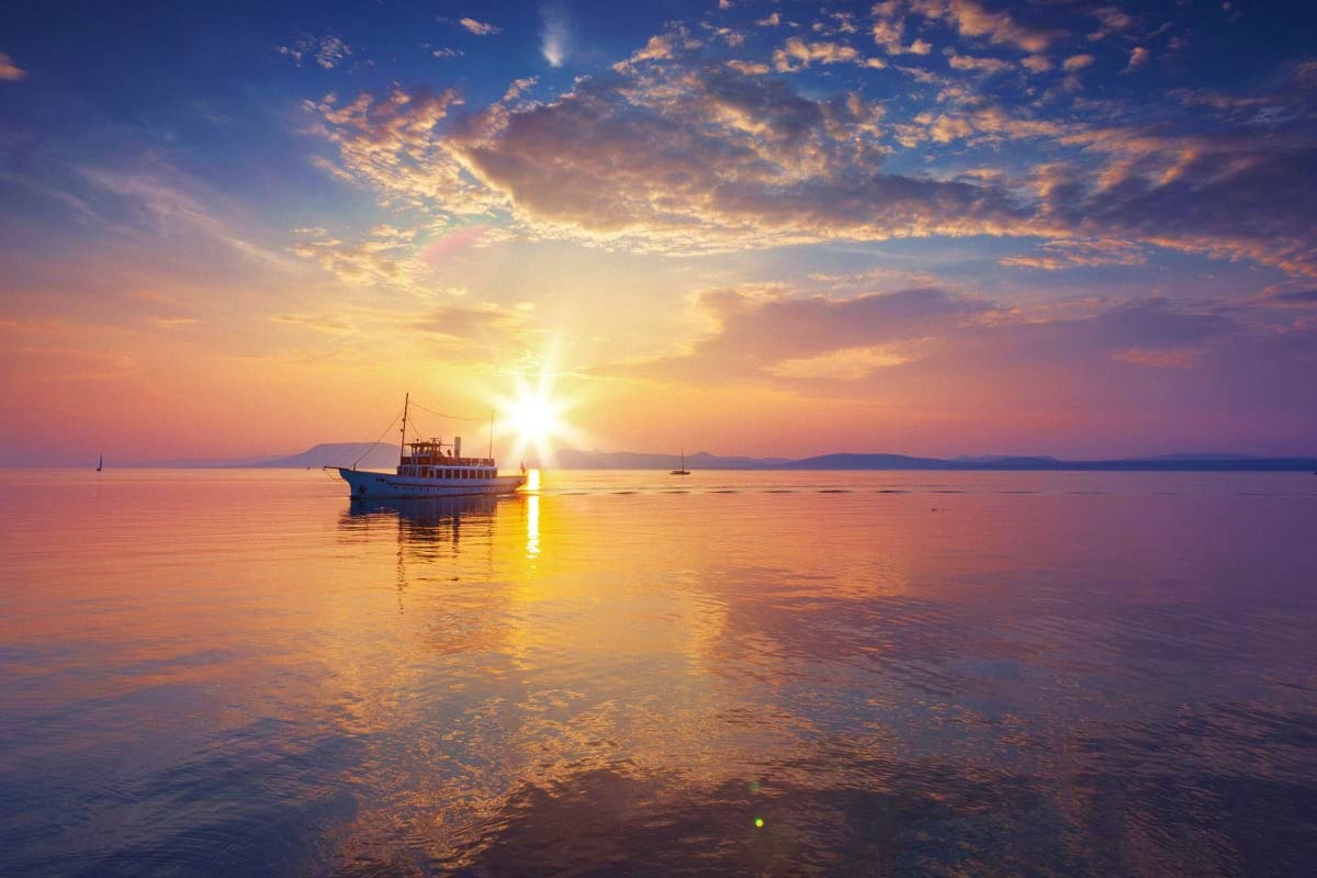 Minsk Super Saver Deal Passenger ferry boat anchored in calm water at golden sunrise with distant mountains and sailboats visible on horizon