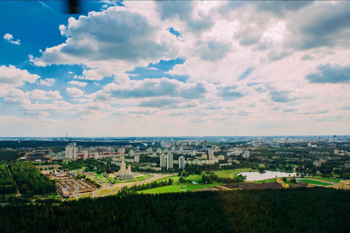 Aerial view of Minsk cityscape with modern buildings, green parks, lakes, and forested areas under cloudy blue sky