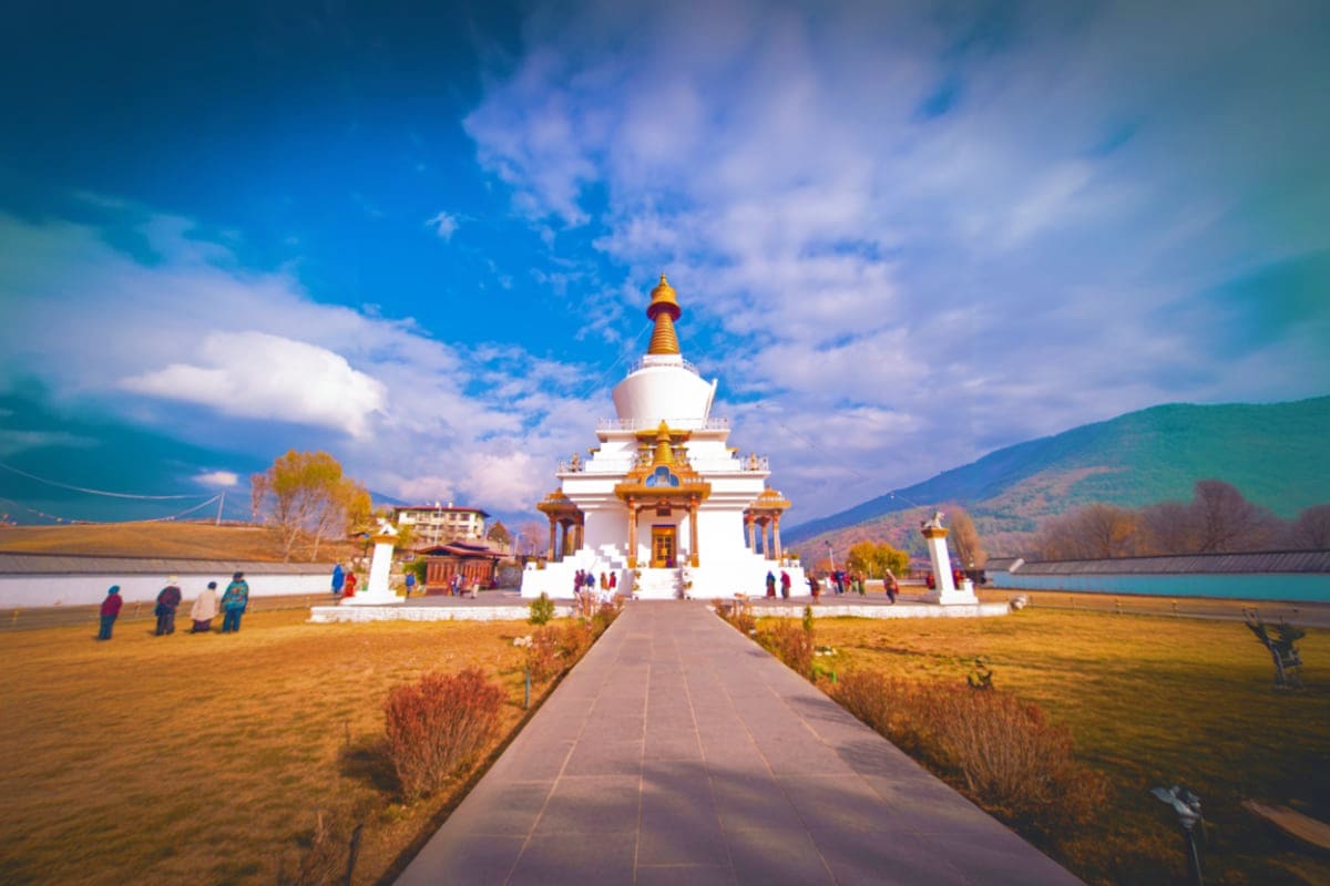Bhutan Short Trip Package with Haa Valley White Buddhist temple with golden spire beside turquoise river, mountains and blue sky, visitors on pathway at Bhutan landmar