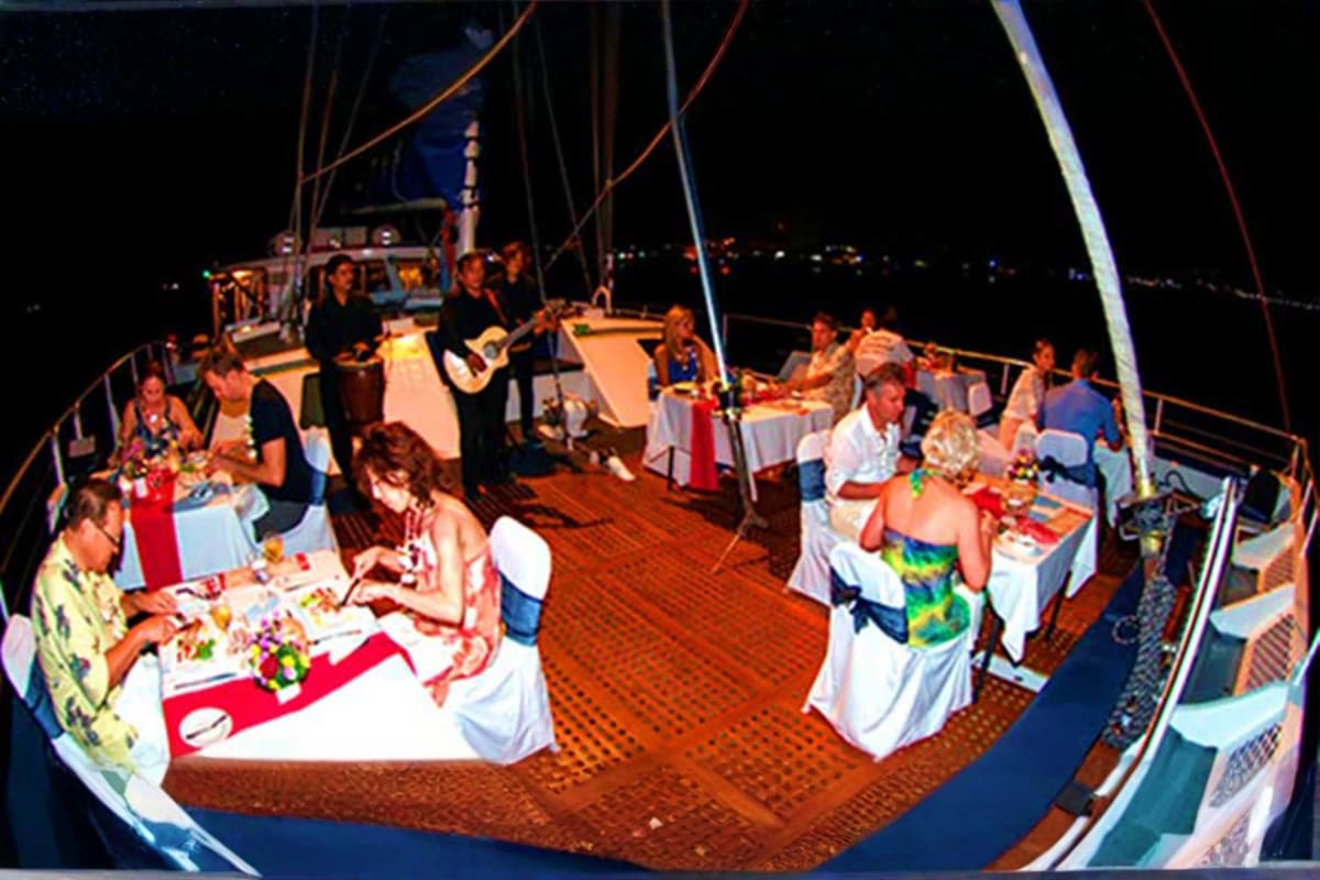 Guests dining at tables on a sailboat deck at night with live music performance and ocean backdrop