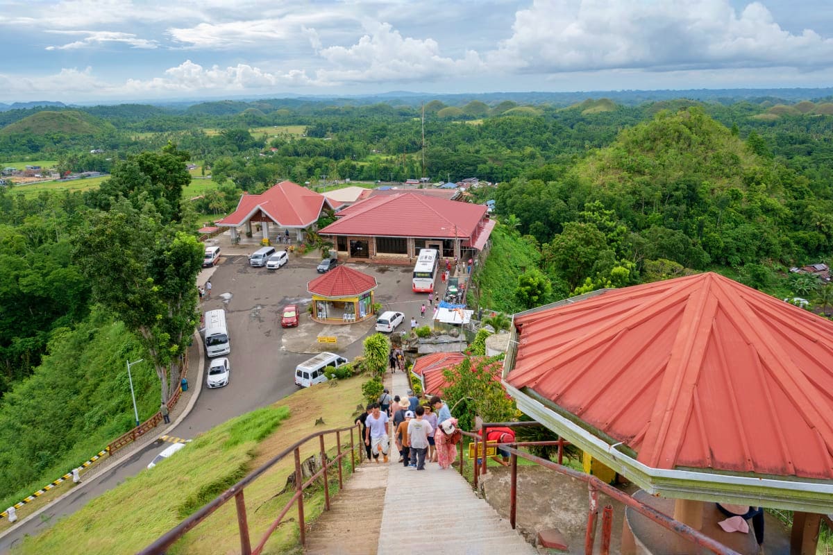 Aerial view of Simply Bohol complex with red-roofed buildings, parking area, and visitors walking down wooden stairs amid lus