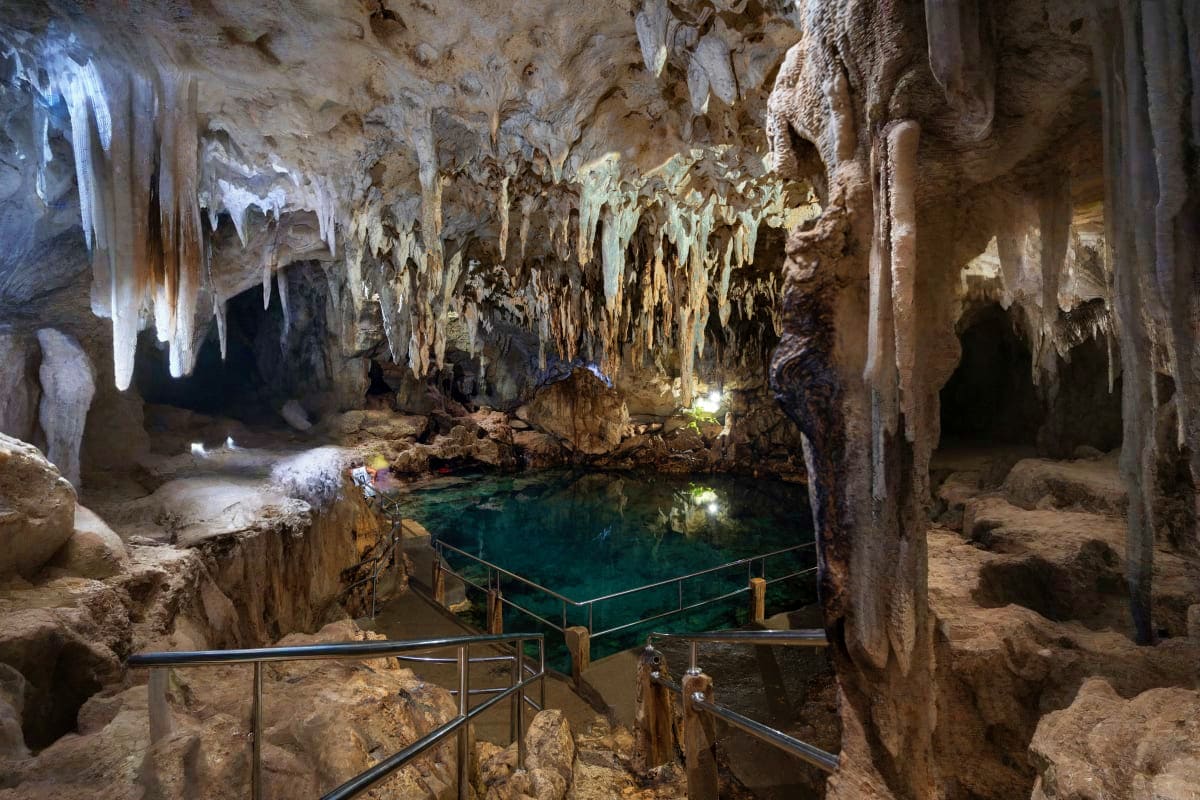 Underground cave with stalactites and stalagmites surrounding a turquoise cenote pool with metal railings for visitor access