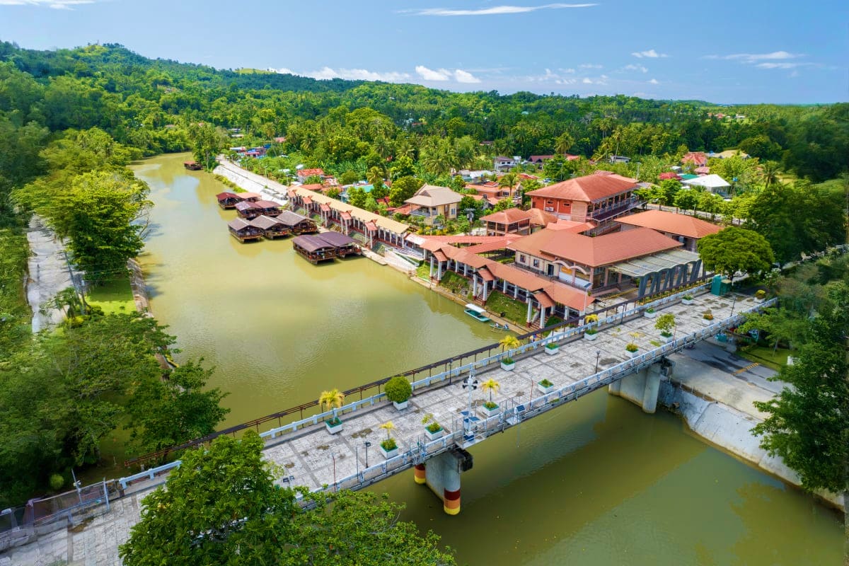 Aerial view of riverside village in Bohol with traditional houses, boats, and bridge spanning calm waterway surrounded by lus