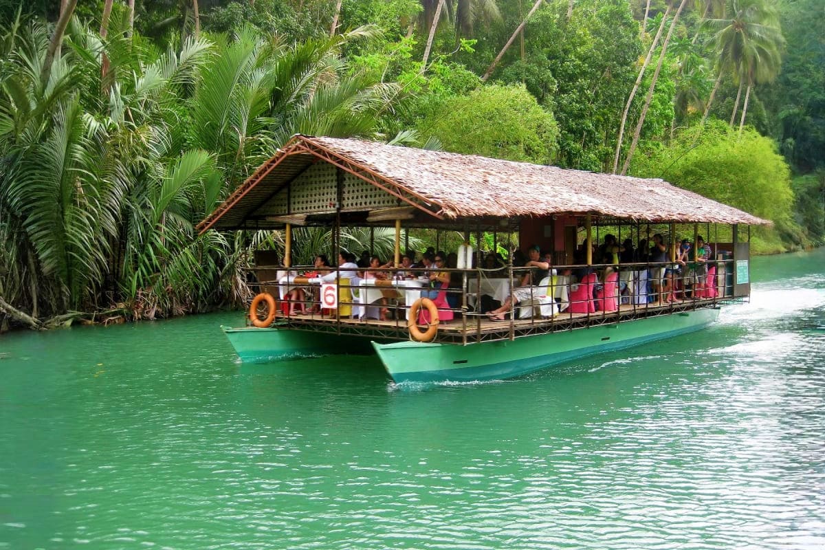 Traditional wooden tour boat with thatched roof carrying tourists on emerald green river surrounded by tropical jungle and pa