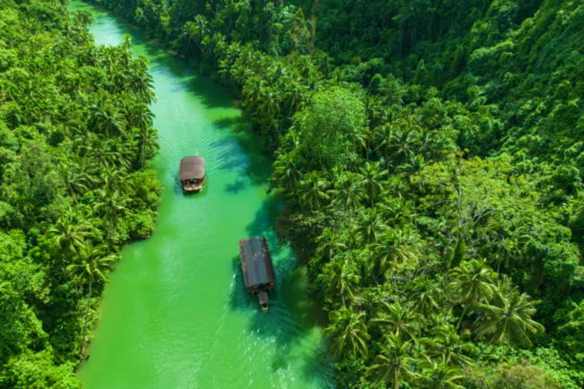 Aerial view of two boats navigating a narrow turquoise river through dense tropical jungle in Bohol, Philippines