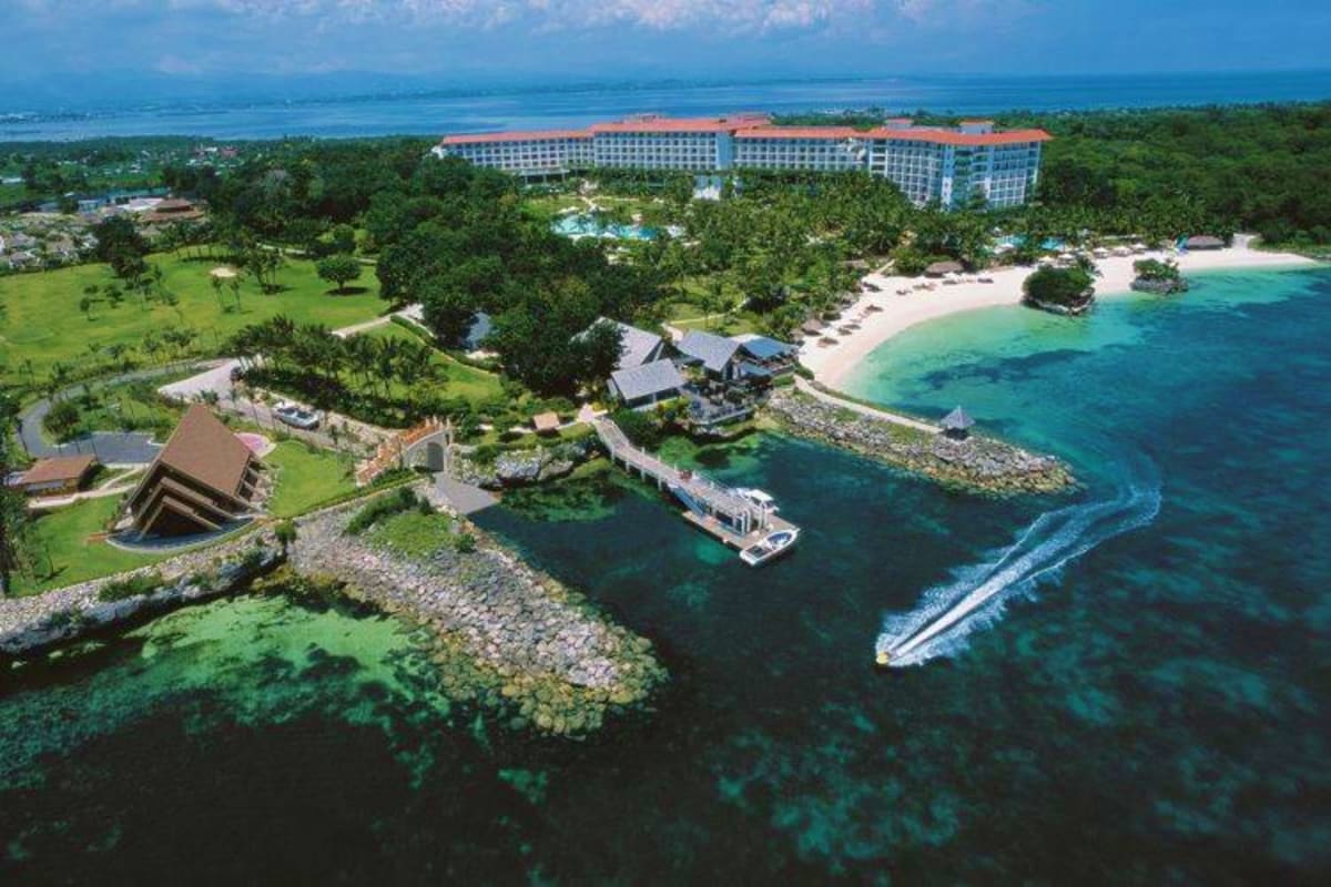 Aerial view of tropical beachfront resort with orange-roofed hotel, white sand beach, turquoise ocean, speedboat, and lush gr