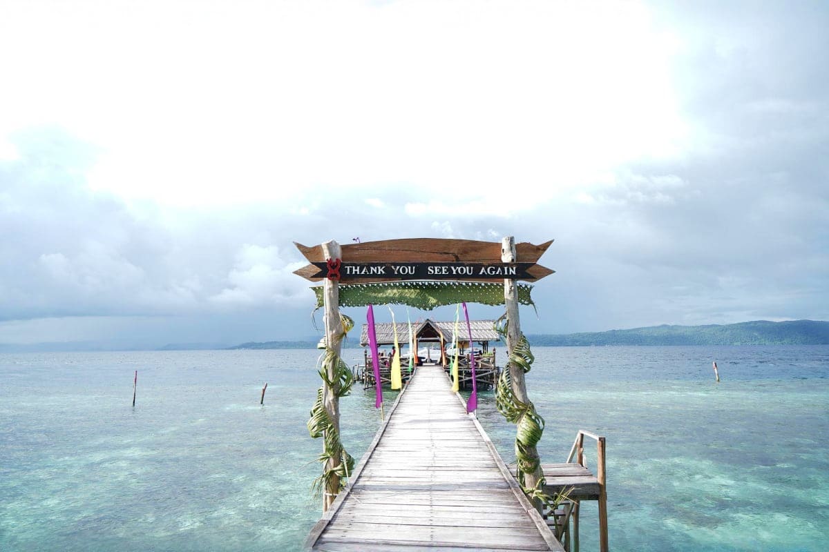 Wooden pier extending into turquoise tropical waters with thank you sign and purple decorations, mountainous coastline in bac