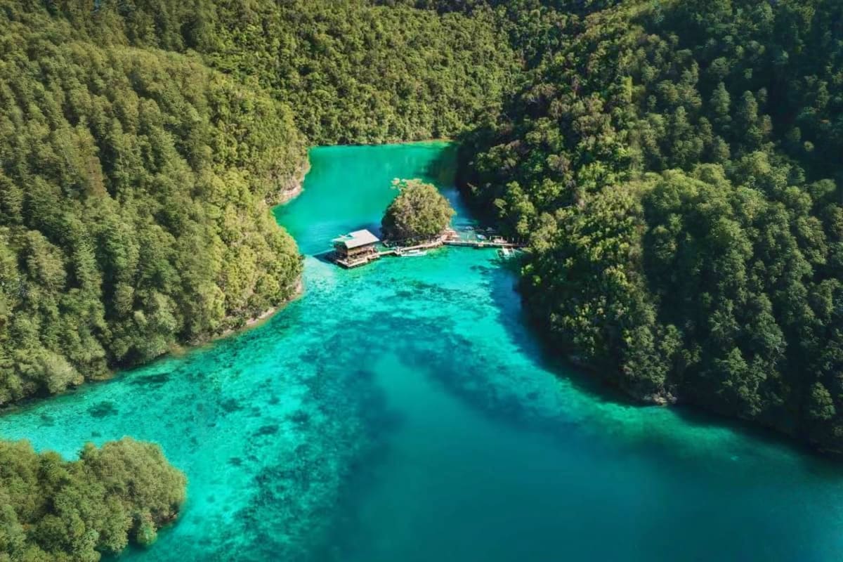 Aerial view of turquoise lagoon surrounded by dense forest with traditional wooden lodge on peninsula in Siargao