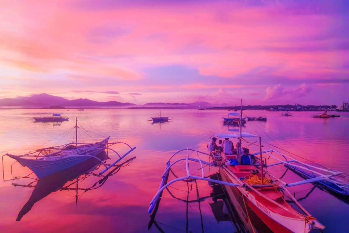 Traditional outrigger boats anchored in calm pink and purple sunrise waters with mountains and other vessels in the backgroun