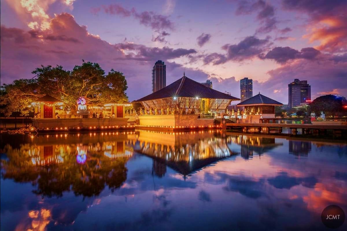 Illuminated Buddhist temple complex with traditional peaked roofs reflecting in calm water at twilight, with modern city skyl