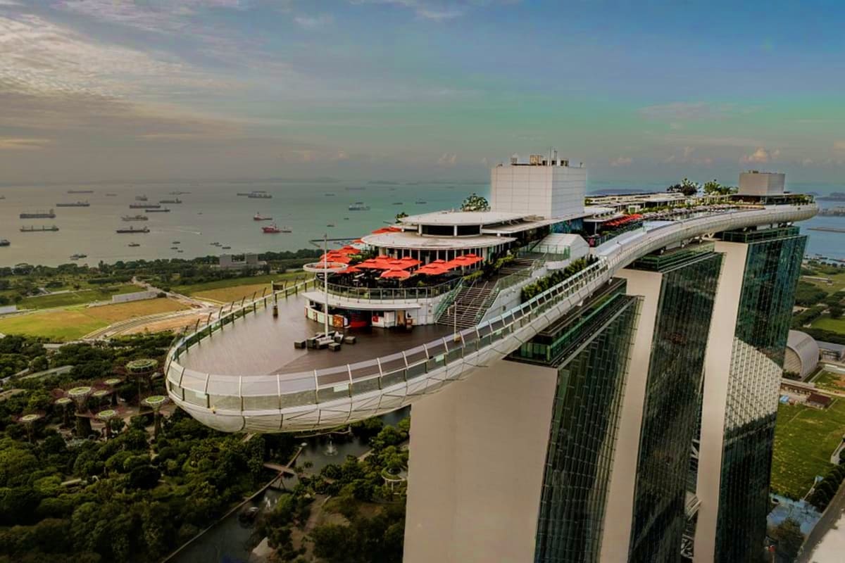 Casino Luxe Aerial view of modern waterfront building with curved walkway, red-roofed restaurant area, and marina with anchored boats in