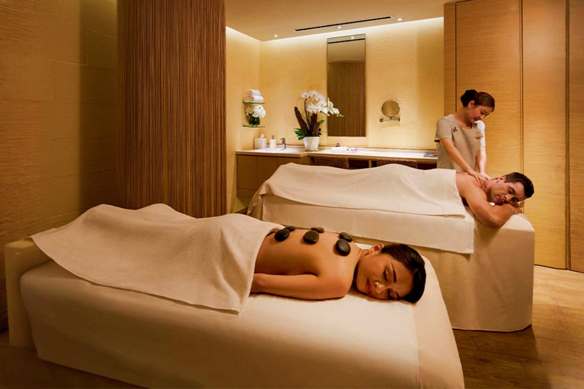 Casino Luxe Spa therapists performing hot stone massage on two clients lying on massage tables in a luxurious treatment room with warm li