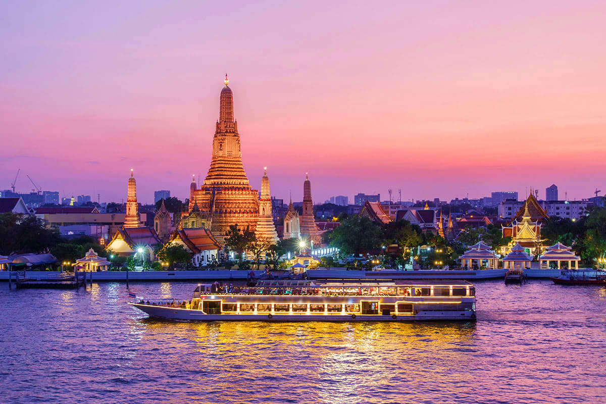 Illuminated golden temple and Buddhist shrines along the Chao Phraya River at sunset with a traditional river cruise boat in 