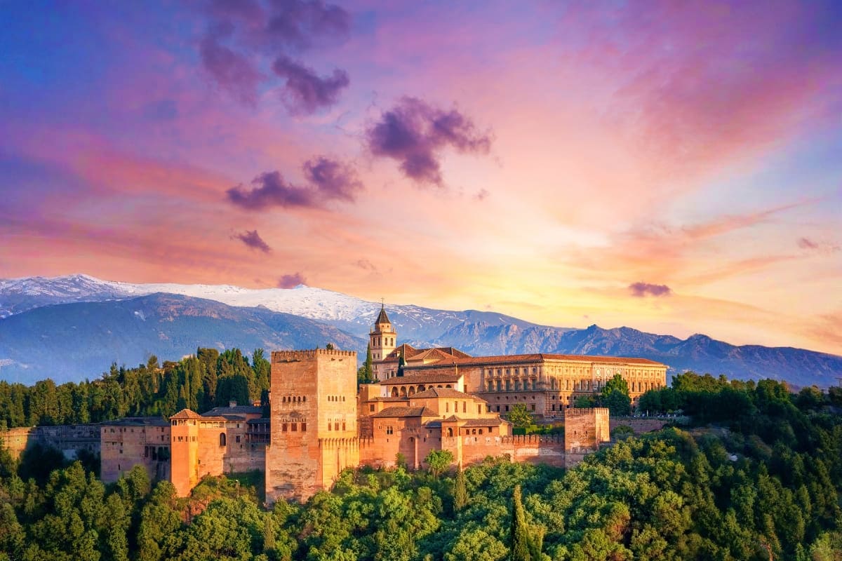 Alhambra palace fortress in Granada at sunset with snow-capped Sierra Nevada mountains and forested hillside in background