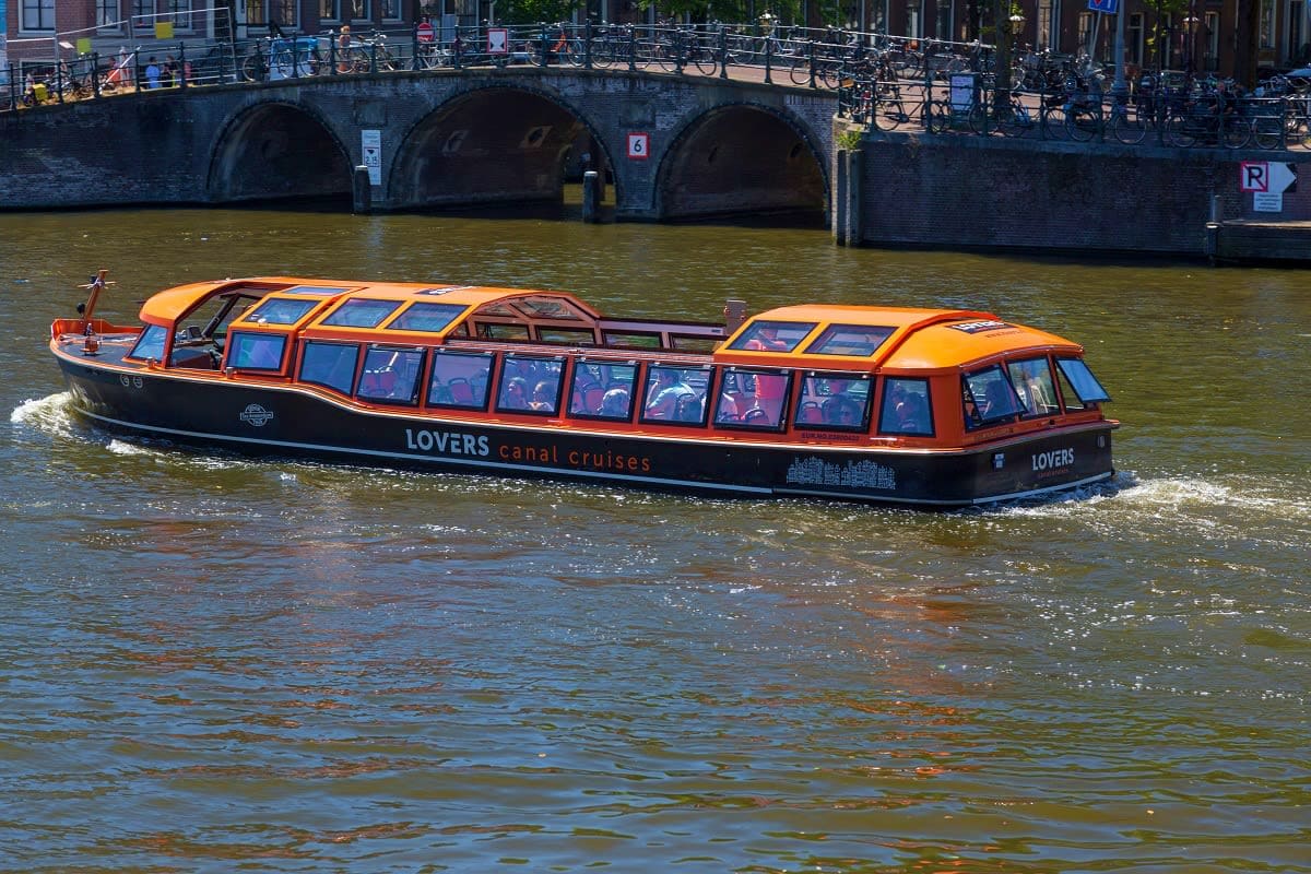 From Brussels to Paris Orange and black Lovers canal cruise boat on Amsterdam canal with passengers, historic stone bridge and bicycles above