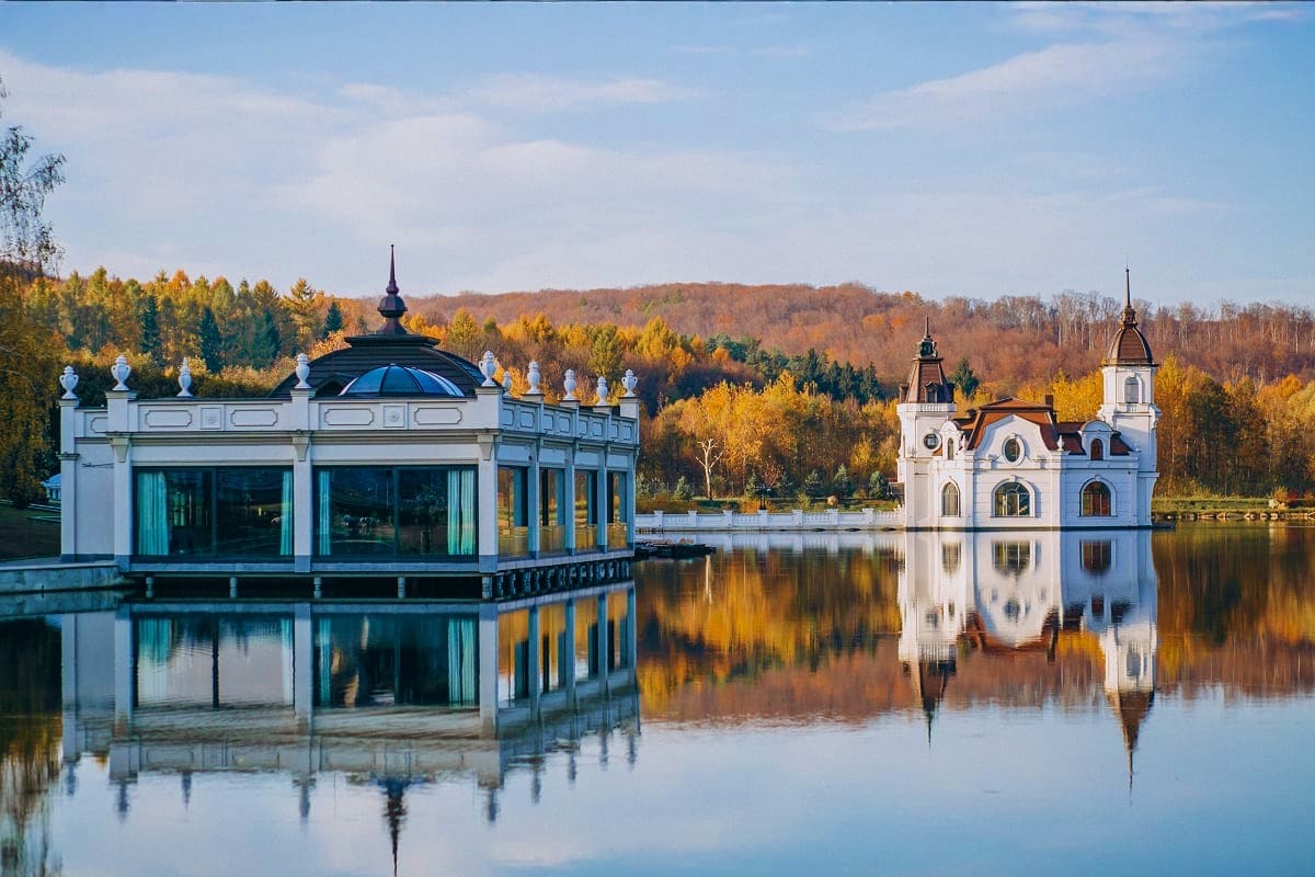 White boathouse and chapel reflected in still lake water surrounded by autumn forest foliage