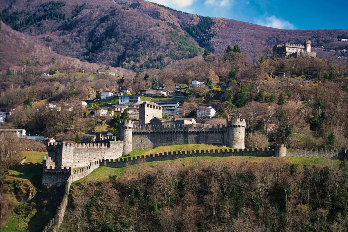 Medieval castle with stone fortifications and towers surrounded by forested mountains and valley settlements in the Alps