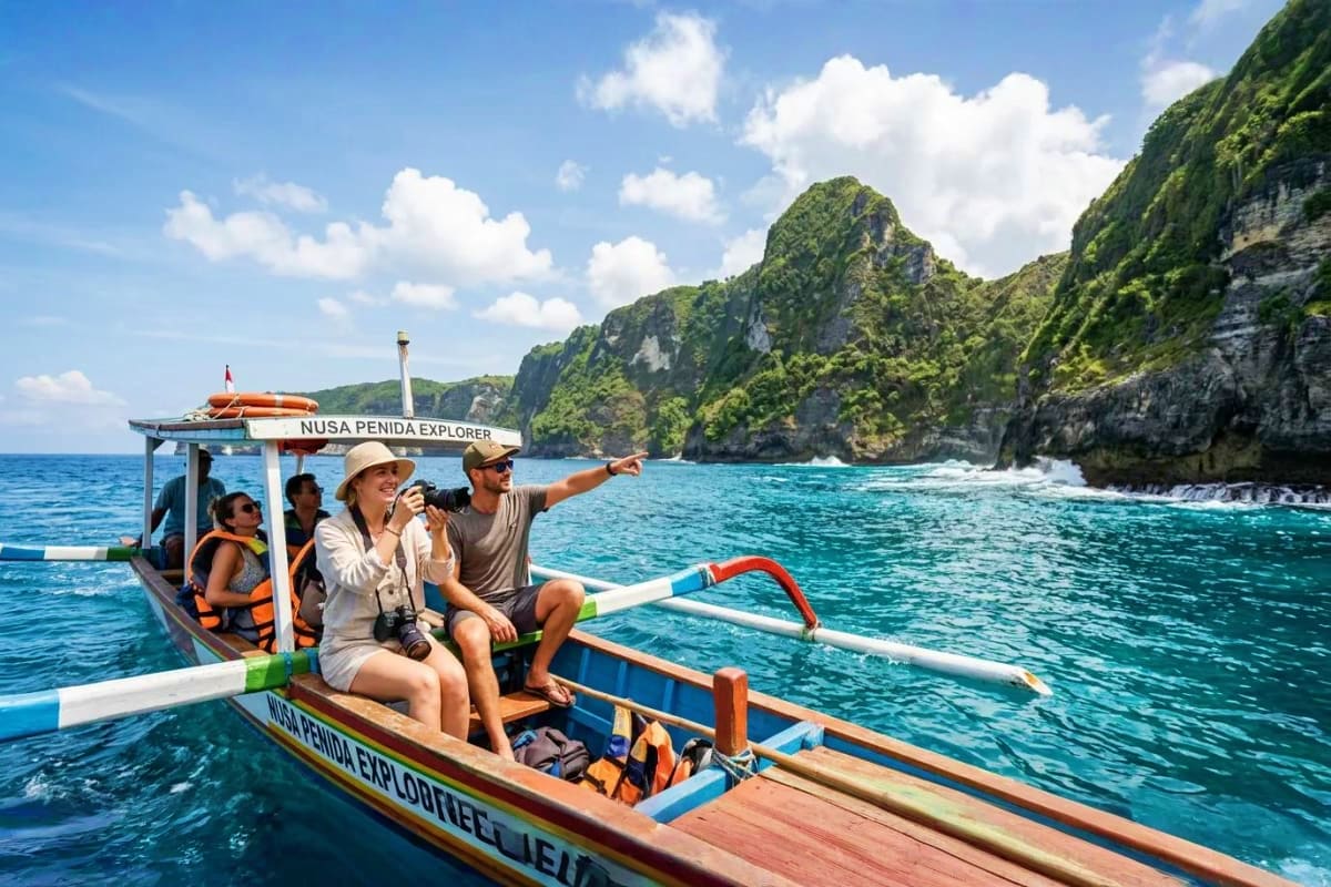 Tourists on traditional wooden boat tour pointing at dramatic green limestone cliffs in turquoise tropical waters, Nusa Penid