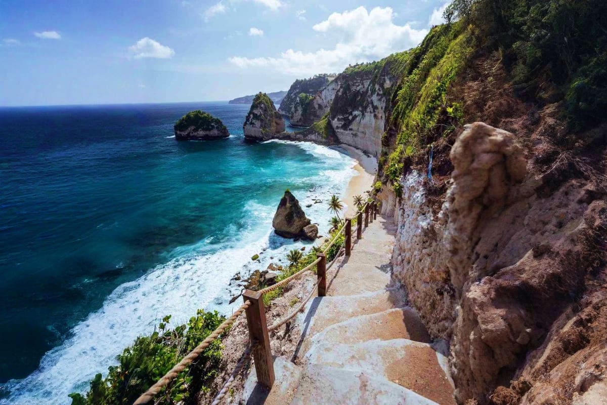 Clifftop pathway overlooking turquoise ocean, dramatic limestone cliffs with lush vegetation, and rock formations in Bali