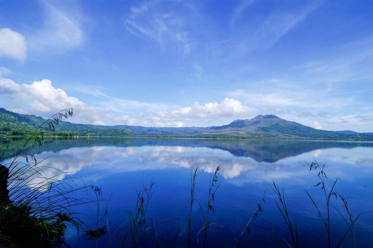 Serene lake landscape with Mount Agung volcano reflected in still blue water, surrounded by lush green mountains and reeds in