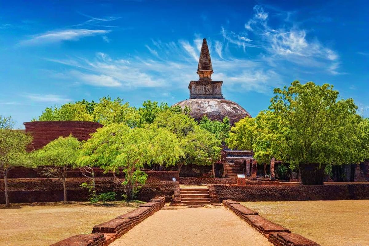 Ancient Buddhist stupa with brick ruins, stone pathway, and lush green trees under blue sky at historical temple complex.