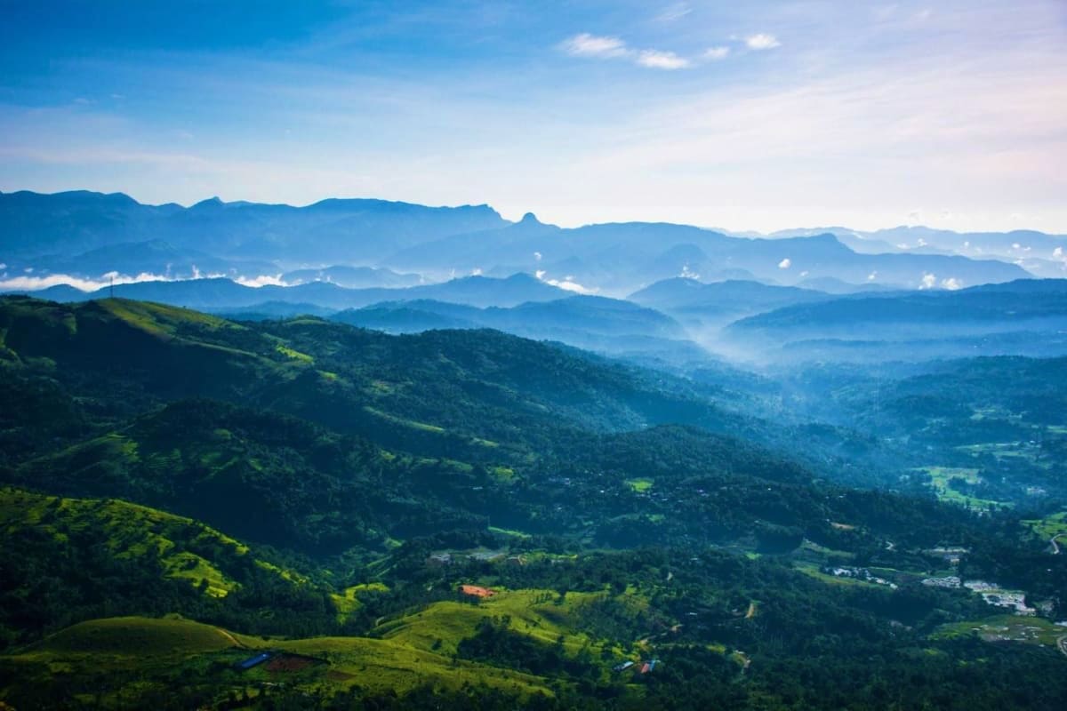 Aerial view of lush green mountains and valleys with layers of blue-tinted mountain ranges in misty distance under clear sky