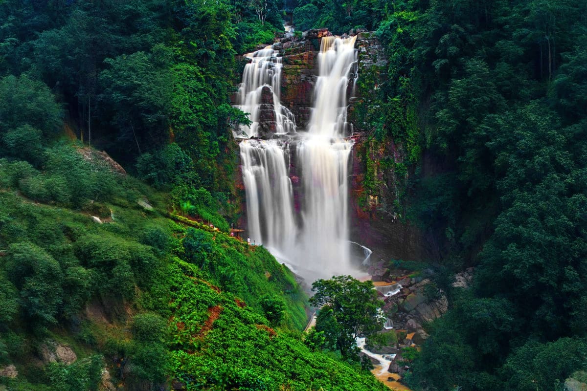 Multi-tiered waterfall cascading through lush green forest with rocky cliffs and stream below