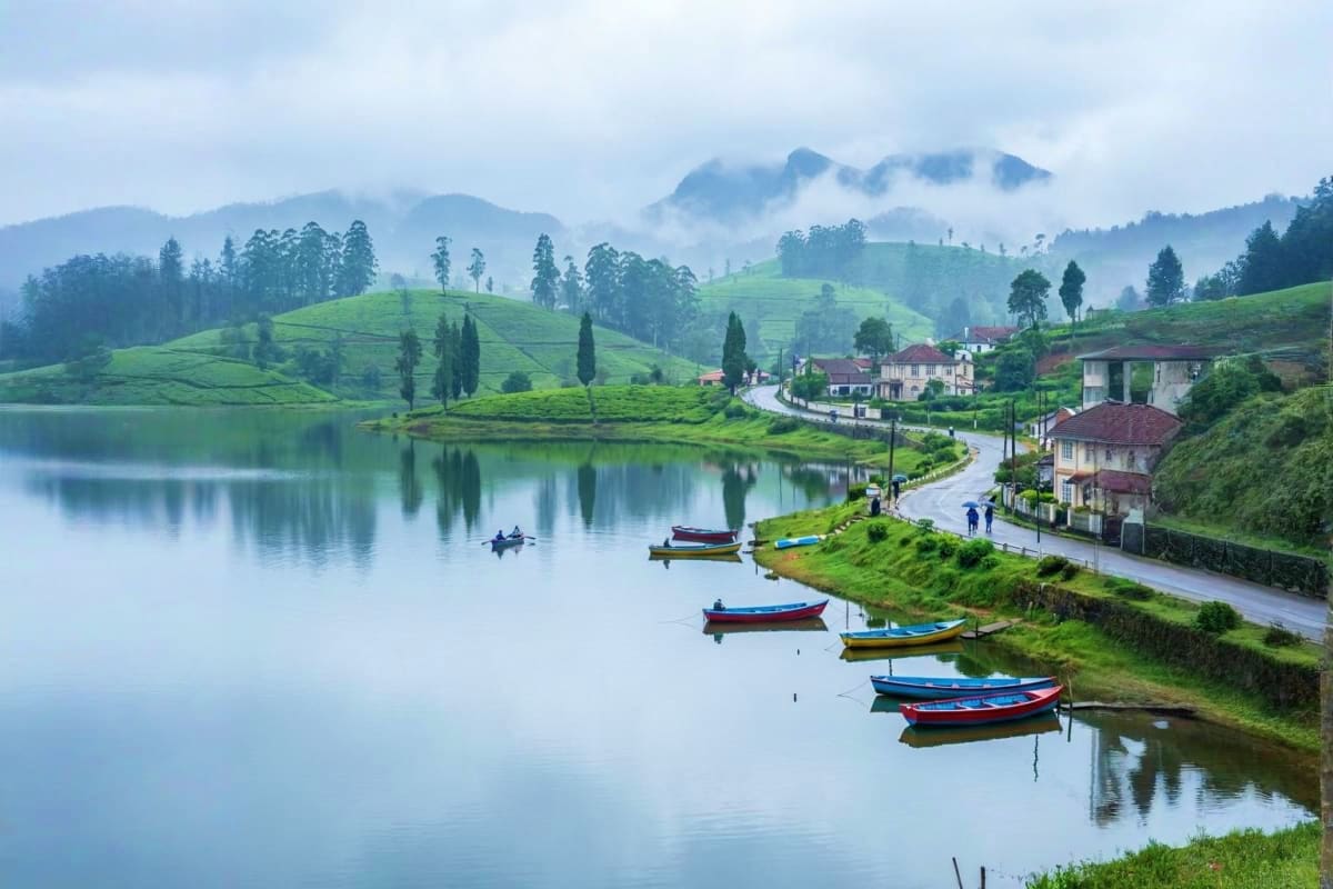 Scenic lakeside village with colorful fishing boats, green rolling hills, cypress trees, and misty mountains reflected in cal