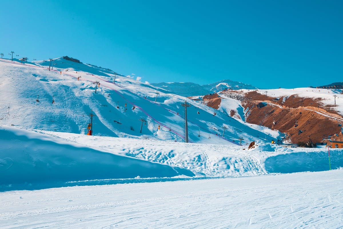 Snow-covered ski slopes with blue runs, ski lifts, and snow-covered mountains under clear blue sky at an alpine ski resort.