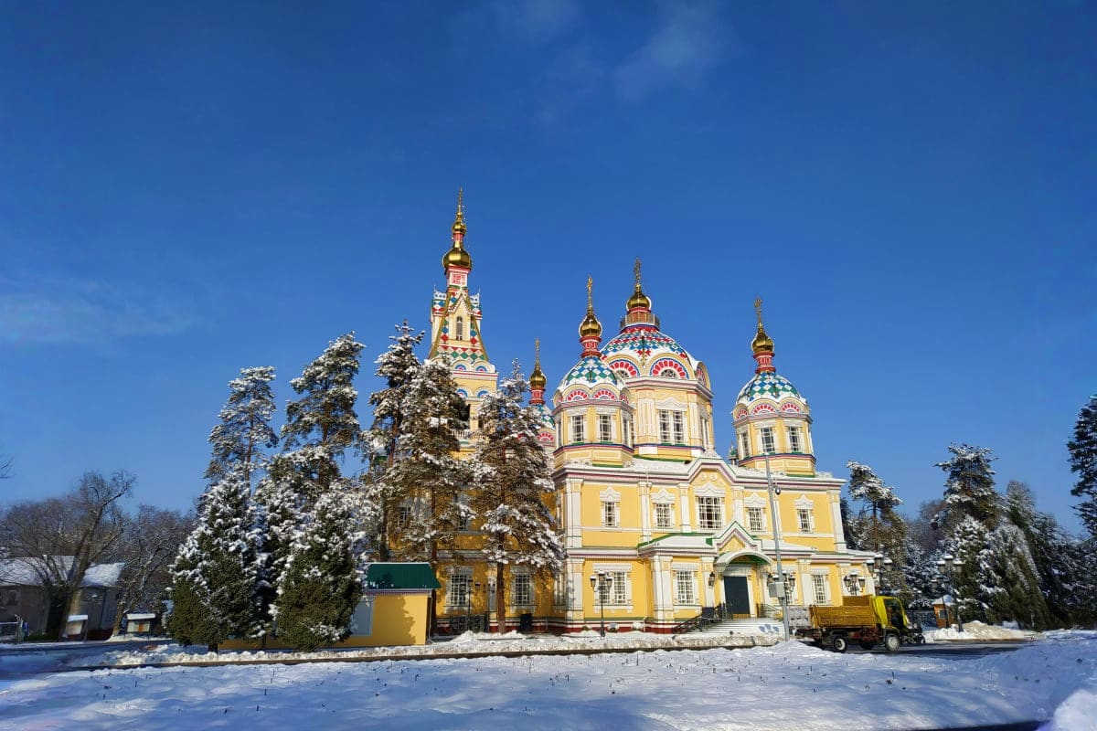 Yellow Orthodox cathedral with golden domes surrounded by snow-covered evergreen trees on a winter day in Kazakhstan