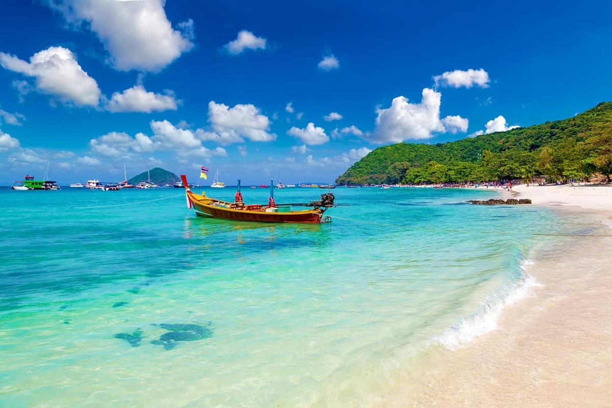 Classic Bangkok and Pattaya Holidays Traditional longtail boat in turquoise waters near sandy beach with forested island and moored boats in Thailand