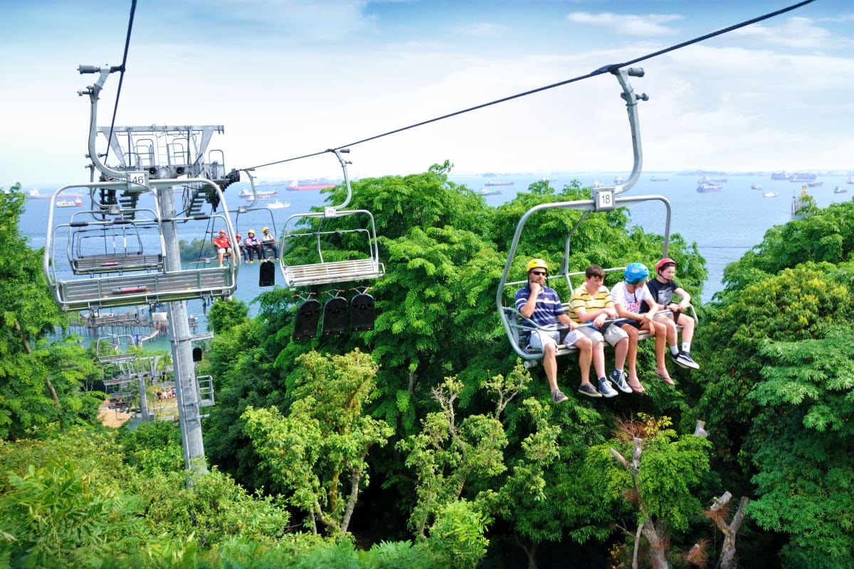 Singapore-Family-Fun-Package Family riding cable car chairlift above lush green forest canopy with Singapore harbor and ships visible in background