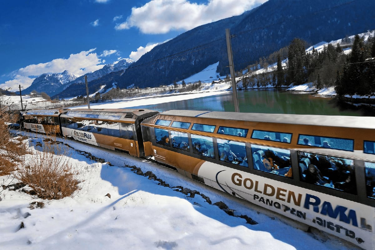 Golden Panorama scenic train docked in snow beside a frozen alpine lake with snow-capped mountains and forested slopes in the