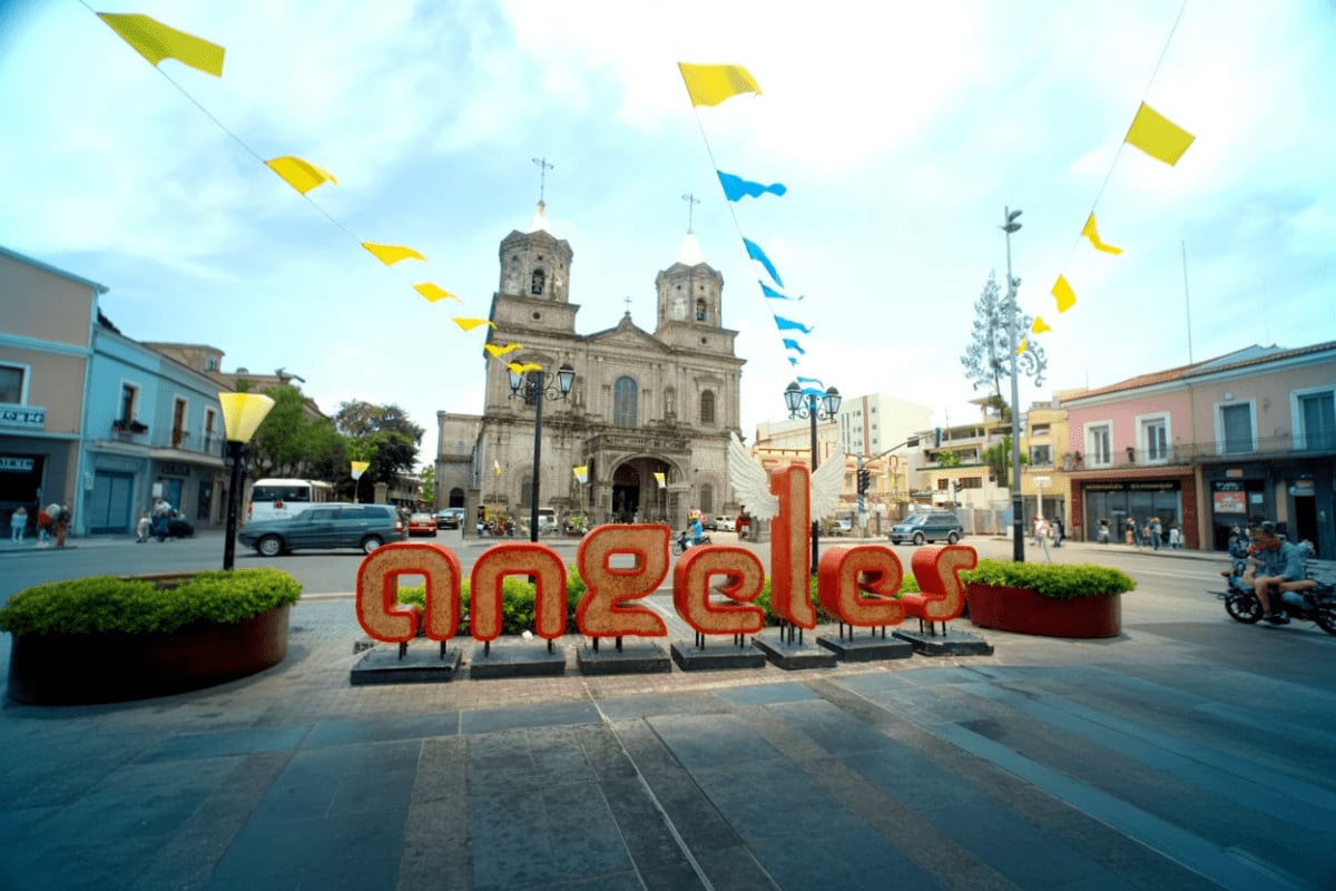 Red "Angeles" sign in historic city plaza with colonial church, yellow and blue flags overhead, and colorful buildings surrou