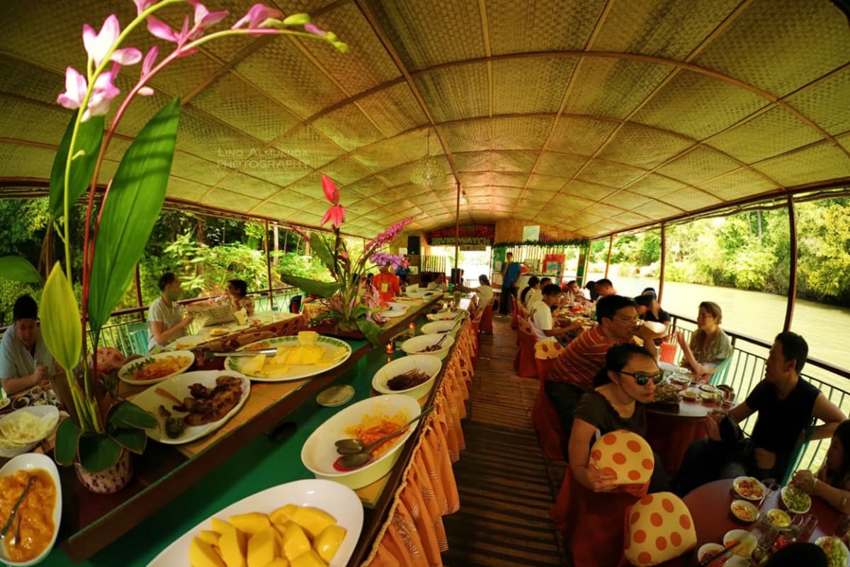 Tourists dining on a covered boat during a Loboc River cruise with tropical flowers and local Filipino dishes served on white