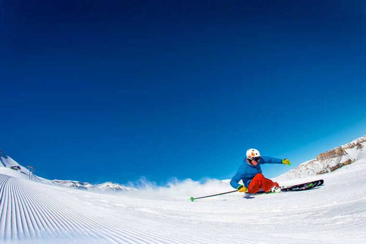 Skier in blue jacket and red pants descending groomed snow slope under clear blue sky with snow-covered mountains in backgrou