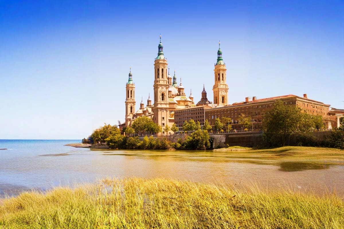 Basilica del Pilar cathedral with golden domes reflected in Ebro River in Zaragoza, Spain under clear blue sky