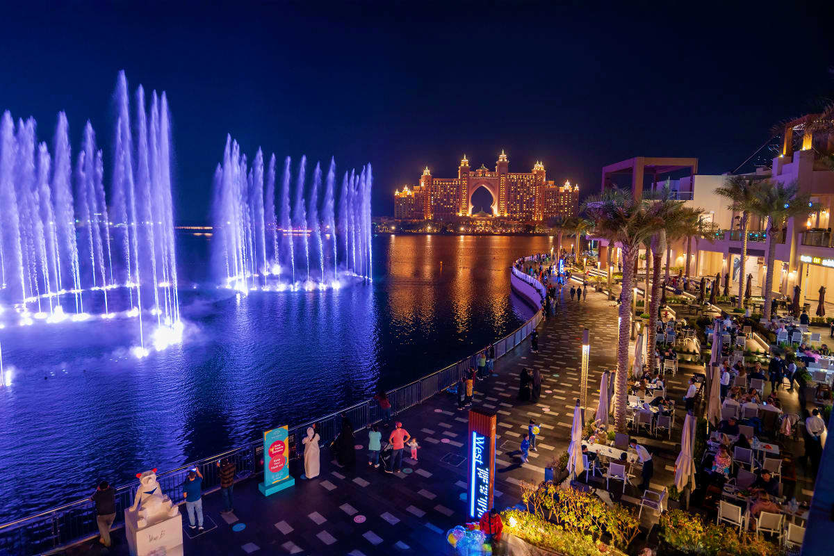 Purple illuminated fountain display at night with Atlantis The Palm hotel lit up across the water and beachfront promenade wi