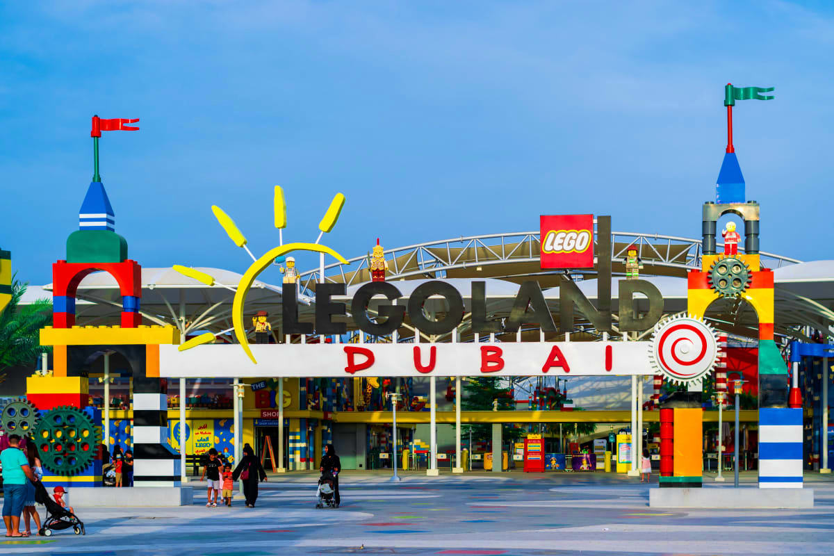 LEGOLAND Dubai entrance with colorful brick facade, yellow ship wheel, red flags, and visitors in the plaza under clear blue 