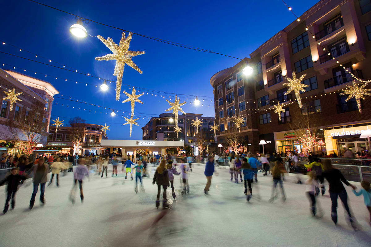 Outdoor ice skating rink at dusk with holiday decorations, golden star ornaments, and crowds of skaters in an urban plaza sur