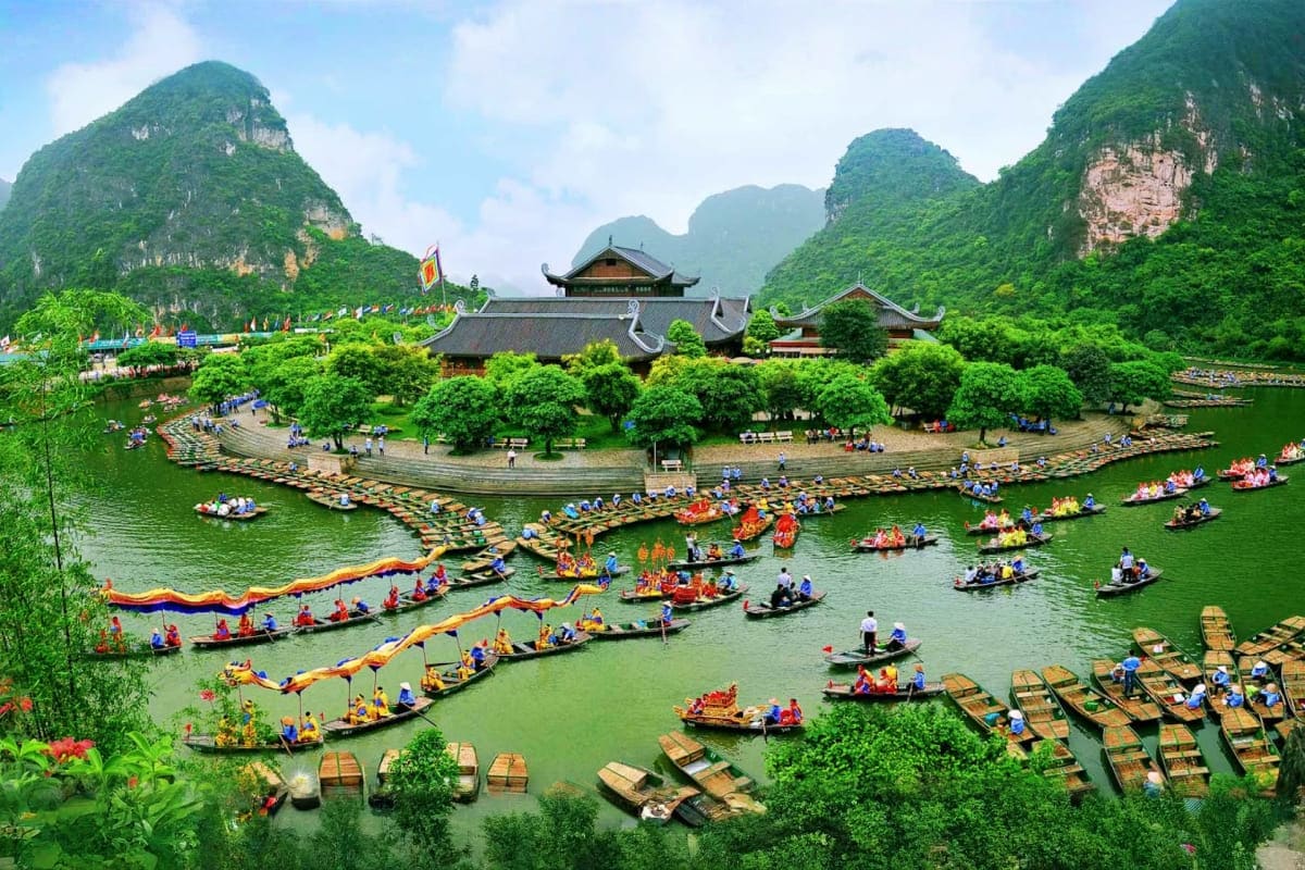 Traditional Vietnamese temple with upturned roofs surrounded by limestone mountains and river filled with ornate boats during