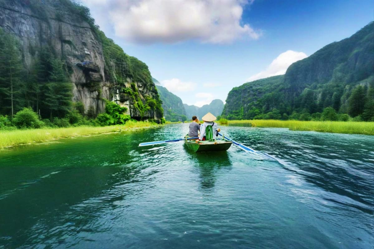 Two people rowing a green boat through calm emerald waters surrounded by towering limestone cliffs and lush green vegetation 