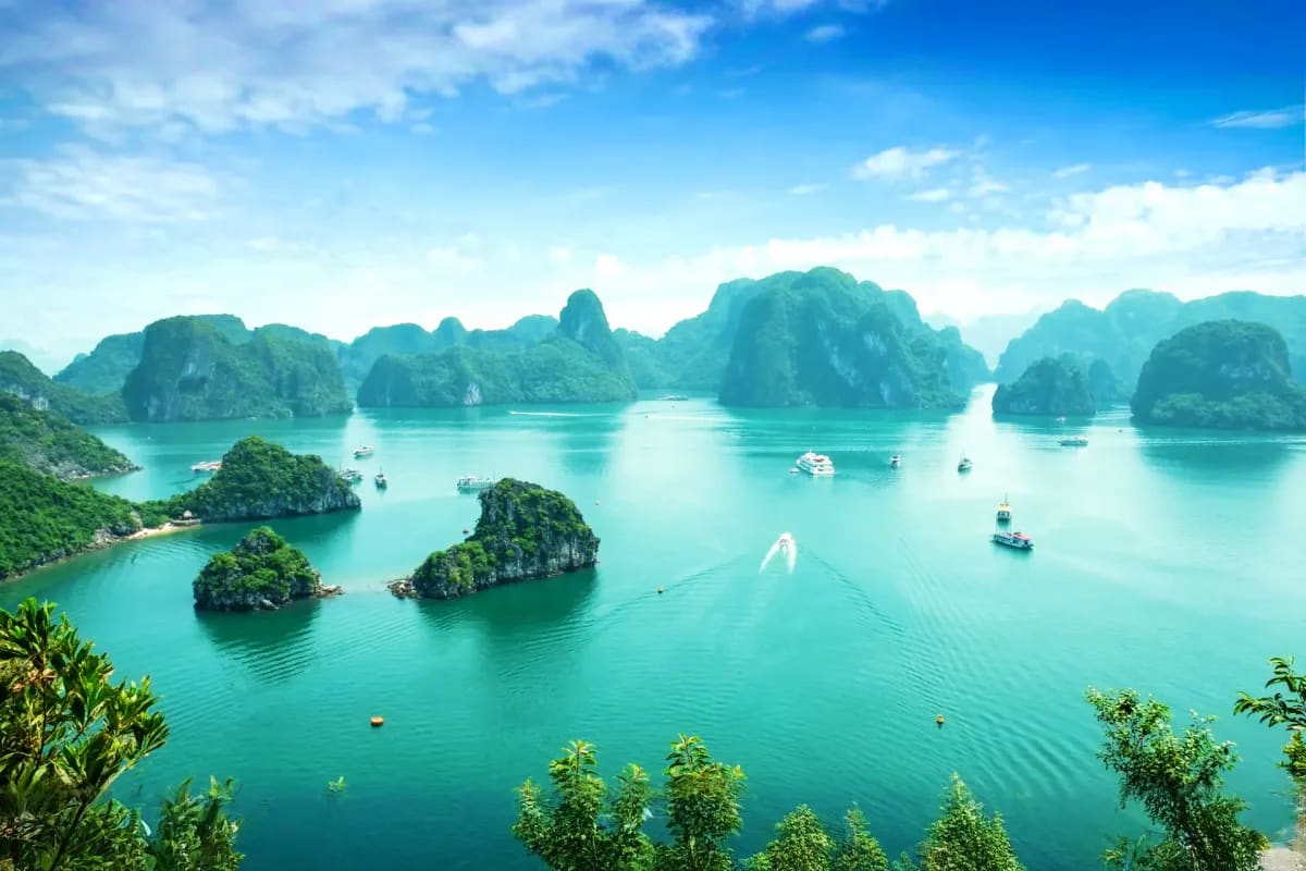 Aerial view of Halong Bay with turquoise water, limestone karst islands, and tourist boats anchored in calm seas under blue s