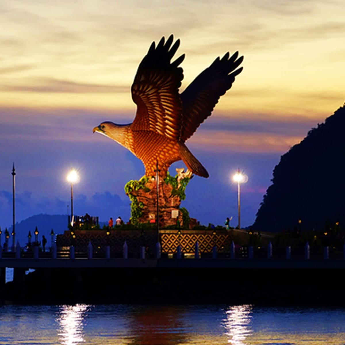 Golden eagle statue at Langkawi waterfront at dusk with illuminated monument and mountain backdrop