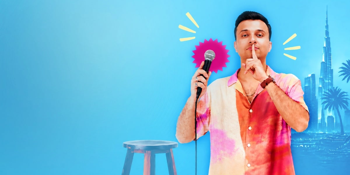 Man in colorful shirt holding microphone making shush gesture with cityscape backdrop and stool
