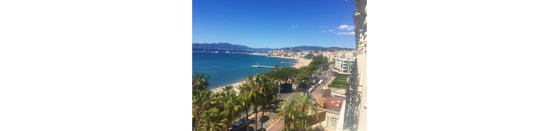 Aerial view of Cannes coastline with palm trees, Mediterranean Sea, sandy beach, and mountains in the distance under clear bl