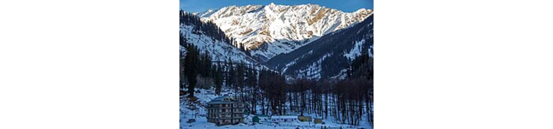 Snow-covered Himalayan valley with pine forests and a building nestled below snow-capped mountain peaks in Manali