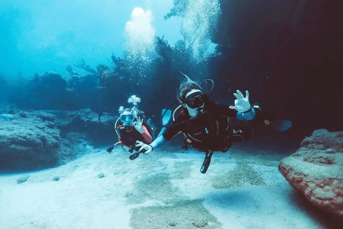 Discover Scuba Diving by WaterColors Two scuba divers exploring underwater reef with sandy bottom and rock formations, sunlight filtering from surface above