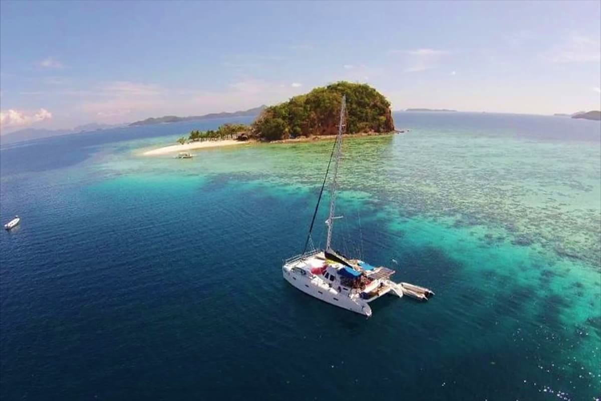 Coron Island Escapade Tour Catamaran sailing near a lush tropical island with white sand beach and turquoise waters in Coron