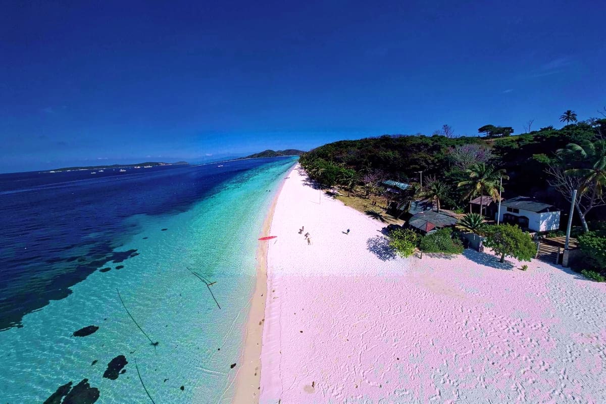 Coron Island Escapade Tour Aerial view of Coron Island's white and pink sand beach with turquoise shallow waters, tropical vegetation, and anchored boat